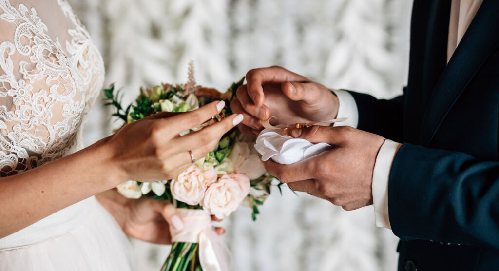 Ein Bräutigam steckt seiner Braut den Ehering an den Finger. Ihre Hände sind mit einem kleinen Strauß weißer Rosen geschmückt.