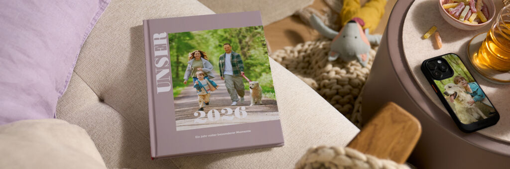 Auf einem Sofa liegt ein lila Fotobuch mit dem Titel „UNSER 2026“ und dem Foto einer Familie mit Hund.